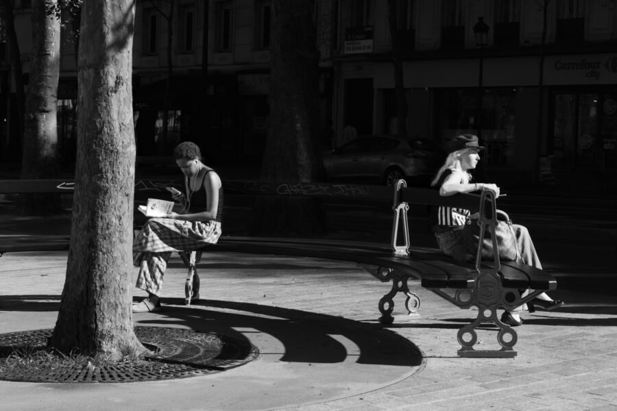 Deux femmes assises sur des bancs à l'ombre des arbres d'une place, l'une lisant, l'autre regardant au loin