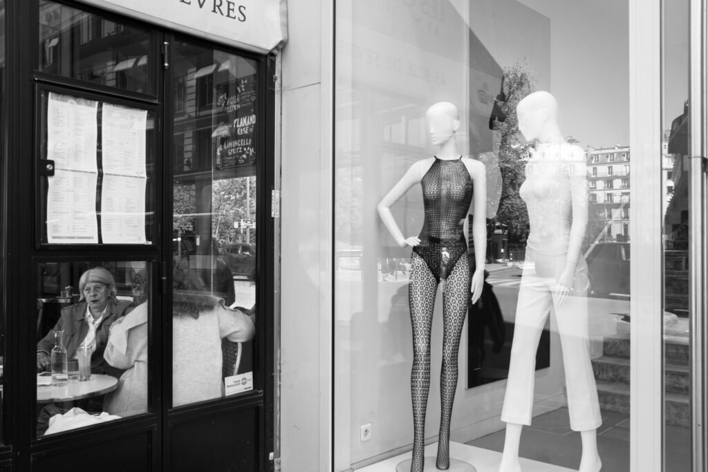 Vitrine de lingerie avec deux mannequins, à côté d'une femme âgée attablée seule derrière la porte vitrée d'un café