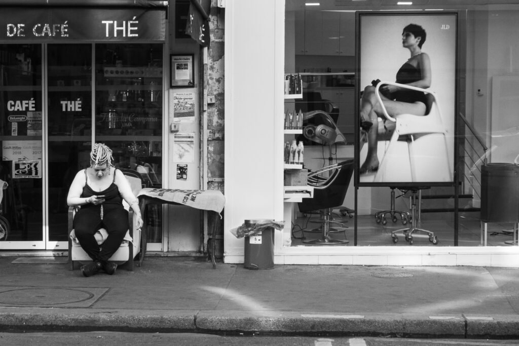 Femme assise seule sur une chaise devant un salon de thé, regardant son téléphone, près d'une affiche de mannequin